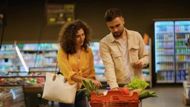 Neşeli genç çift marketten taze sebze ve meyve seçiyor. Kadın ürünleri seçerken erkek alışveriş listesini kontrol ediyor.