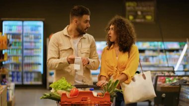 Genç aile, süpermarkette uzun bir market fişini kontrol ederken şok olmuş ve stresli hissediyor. Çift yüksek gıda fiyatlarını tartışıyor