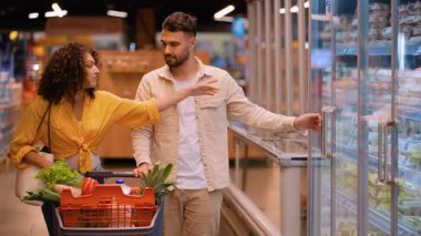 Genç çift süpermarkette dondurulmuş bir vitrinden yumurta seçiyor. Adam bir karton alıyor ve el arabalarıyla alışverişe devam ediyorlar.