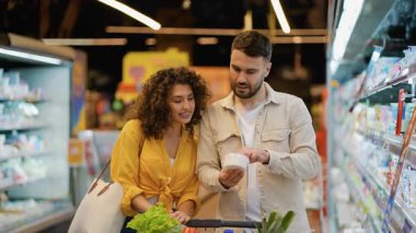 Mutlu genç çift marketteki soğutulmuş raflardan süt ürünlerini tartışıyor ve seçiyor. Kadın ve erkek birlikte gülümsüyor.