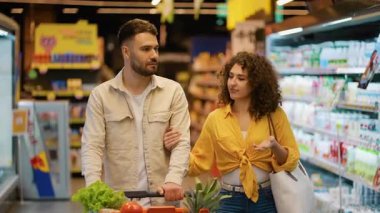 Alışveriş yaparken süpermarket koridorunda yürüyen neşeli bir çift. Kadın mutlu bir şekilde sohbet ederken adam sebzeli bir arabayı itiyor.
