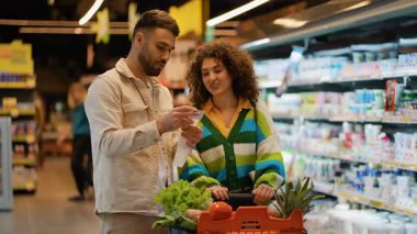 Markette taze ürün alırken alışveriş listelerini kontrol eden genç çift sağlıklı yiyecekleri birlikte seçiyorlar.