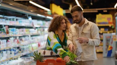 Genç mutlu çift süpermarkette birlikte alışveriş yapıyor. Elinde alışveriş listesi olan bir adam ve sağlıklı beslenme için taze sebze seçen bir kadın.