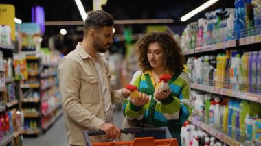 Genç mutlu çift süpermarkette birlikte alışveriş yapıyor, raftan temizlik ürünlerini seçiyor ve onları alışveriş arabasına koyuyor.