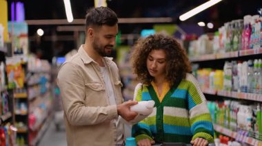 Genç evli çift süpermarket koridorunda temizlik ürünü şişesinin etiketini okuyor. Karı koca alışveriş listelerini tartışıyor.