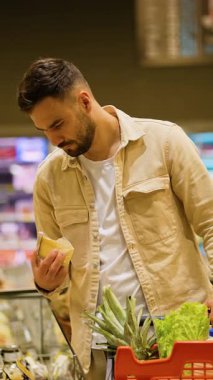 Bakkalda soğutulmuş bir vitrinden peynir seçen odaklanmış genç bir adam. Onun alışveriş sepetinde taze marul ve bir ananas var.