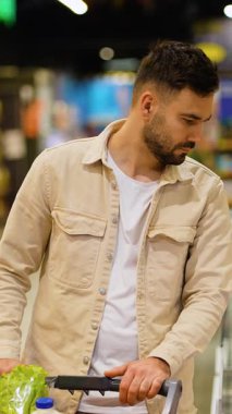 Odaklanmış sakallı genç adam alışveriş arabasını itiyor ve marketin koridorundaki raftan salam sosisi seçiyor.