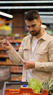 Genç beyaz bir adam alışveriş arabasıyla modern bir süpermarkette taze yiyecek alırken alışveriş listesini dikkatle kontrol ediyor.