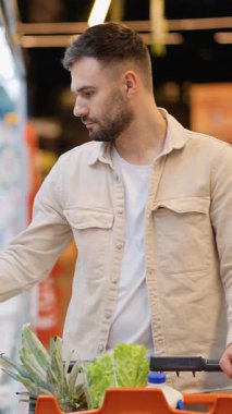 Sakallı odaklanmış genç bir adam markette alışveriş yaparken bir ürünün etiketini dikkatle okur.