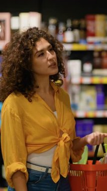 Güzel kıvırcık saçlı kadın modern bir süpermarket koridorunda alışveriş yaparken bir şişe gül şarabını dikkatlice seçiyor.