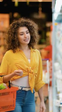 Güzel kıvırcık saçlı kadın market alışverişi yapıyor. Gülümseyen müşteri bir sepet tutuyor ve raftan taze süt ürünleri seçiyor.