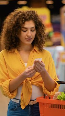 Güzel kıvırcık saçlı kadın alışveriş listesini kontrol ederken bir marketin koridorunda elinde sepetle ürünleri dikkatlice seçiyor.