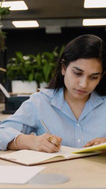 Odaklanmış Hintli kız öğrenci özenle bir dergiye notlar yazıyor. Modern bir çalışma alanında masa başında oturan genç profesyonel bir kadın.
