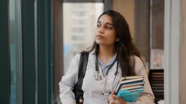 Beyaz önlüklü genç Hintli tıp öğrencisi, elinde kitaplarla üniversite koridorunda yürüyor ve kameraya gülümsüyor.