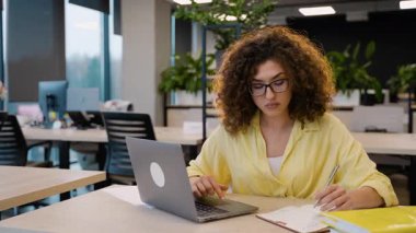 Kendini derslerine odaklamış kıvırcık saçlı ve gözlüklü bir kız öğrenci modern bir üniversite kütüphanesinde deftere notlar yazıyor. Modern bir kütüphanede online öğrenim görüyor.