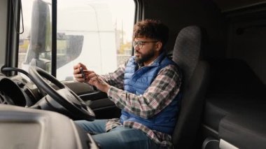 Kamyon kabininde gözlüklü ciddi bir kamyon şoförü oturuyor ve iletişim ve navigasyon için akıllı telefon kullanıyor.