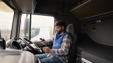 Gözlüklü genç sakallı adam bir tırın şoför koltuğunda oturuyor. Erkek sürücü mola veriyor ve telefon görüşmesi yapıyor.