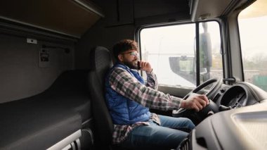 Kamyon kabininde direksiyonda oturan profesyonel erkek sürücü telefonla konuşuyor ve akıllı telefonunu kontrol ediyor.
