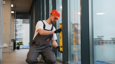 Yeni bir pencere kurulumunu kontrol etmek için ruh seviyesinde bir araç kullanan sert şapkalı genç bir erkek yükleyici. Profesyonel tamirci kesinliği garanti ediyor.