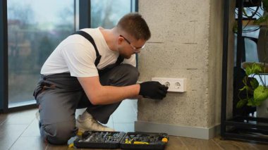 Güvenlik gözlüğü takan ve üniforma giyen genç bir elektrikçi modern ofisin beton duvarına yeni bir üçlü elektrik prizi takıyor.