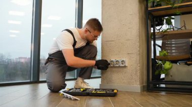 Üniformalı genç bir adam ofis binasına yeni bir elektrik prizi kuruyor ve monte ediyor. Elektrikçi elektrik prizini inceliyor.