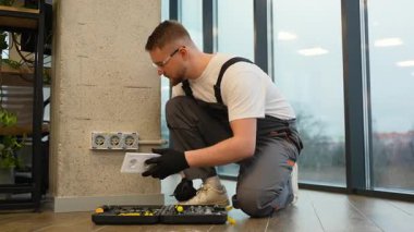 İş üniformalı ve güvenlik gözlüklü genç elektrikçi modern bir ofiste beton bir duvara yeni bir elektrik prizi yerleştiriyor.