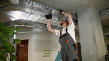 Profesyonel erkek elektrikçi, modern ofis içinde kablosuz matkap kullanarak kablo tepsisine bağlantı kutusu yerleştiriyor.
