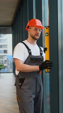 Profesyonel işçi tulumu ve şapkalı bir tamirci ruh seviyesiyle duvarı kontrol ediyor. Yenileme sırasında dikey hizalamayı sağlayan yapıcı