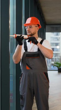 Sert şapkalı ve tulumlu profesyonel tamirci pencere kurulumu ya da tamirat için silikon dolgulu bir kazan tabancası hazırlıyor.