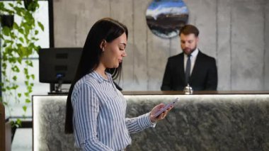 Lüks bir otele giriş yapan genç bir bayan kayıt için telefonunu kullanırken arka planda bir erkek resepsiyonist bekliyor.