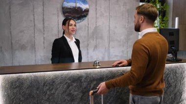 Dost canlısı bir bayan resepsiyonist, erkek bir konuğa kontrol işleminde yardımcı oluyor. Adam bavulla gelir ve gülümseyen tezgahtardan bir anahtar kartı alır.