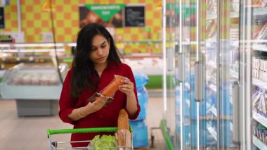 Genç Hintli kadın marketteki alışveriş arabasına koymadan önce yiyecek paketinin etiketini dikkatlice okuyor.