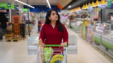 Güzel genç bir kadın, içi taze sebze ve sebze dolu bir alışveriş arabasını itiyor. Bir süpermarketin koridorlarında yürüyor.