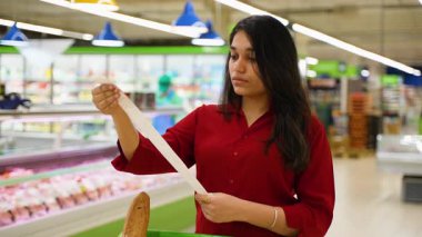 Endişeli genç bayan market alışverişinden sonra uzun ve pahalı bir faturayı dikkatle kontrol ediyor. Yüksek fiyatlar nedeniyle şok olmuş müşteri tasarıyı inceliyor