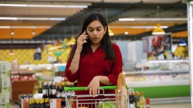 Akıllı telefonuyla konuşan genç Hintli bir kadın markette raflarla dolu bir alışveriş arabasını iterken...