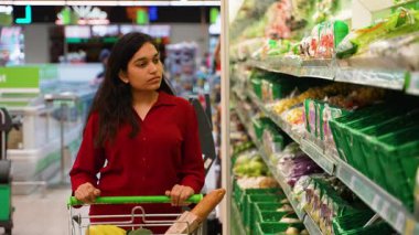 Genç bir kadın alışveriş arabasını itiyor ve raftan taze marul seçiyor. Sağlıklı gıda seçenekleri ve süpermarketten market alışverişi