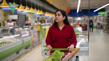 Güzel genç bir kadın, sebze ve ekmek de dahil olmak üzere taze yiyecek dolu bir alışveriş arabasını modern bir süpermarket koridorunda itiyor.