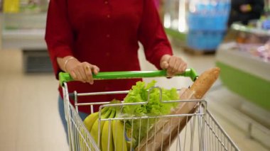 Kırmızı tişört giyen tanınmayan bir kadın marketin koridorunda sağlıklı yiyecek dolu bir alışveriş arabasını itip günlük alışverişini yapıyor.