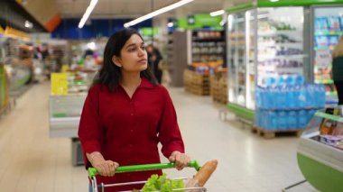 Genç Hintli kadın elinde alışveriş arabasıyla modern bir markette yürüyor. Müşteri sağlıklı bir yemek için taze ürünler seçiyor.