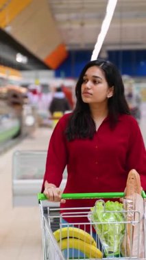 Kırmızı tişörtlü genç, güzel Hintli kadın, sağlıklı yiyeceklerle dolu alışveriş arabasını modern bir süpermarket koridorunda azimle itiyor.