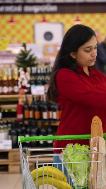 Kırmızı tişörtlü düşünceli bir Hintli kadın market alışverişi yapıyor. Sepetine koymadan önce süt şişesindeki etiketi dikkatlice okuyor.