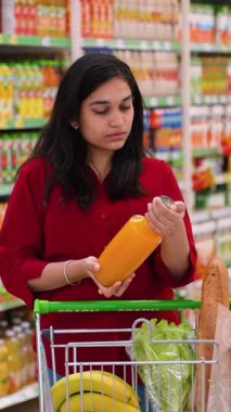 Düşünceli Hintli kadın meyve suyu şişesini inceliyor, marketteki alışveriş arabasına koymadan önce etiketi okuyor.