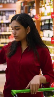 Genç Hintli kadın marketin alkol reyonunda beyaz şarap şişesinin etiketini dikkatlice okuyor.