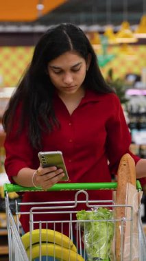 Genç Hintli kadın alışveriş listesini akıllı telefondan kontrol ederken marketin koridorunda içi temiz yiyeceklerle dolu bir market arabasına yaslanıyor.
