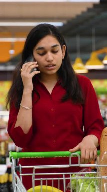 Güzel, genç Hintli bir kadın markette alışveriş yaparken akıllı telefonuyla konuşuyor, alışveriş arabasıyla bekliyor.