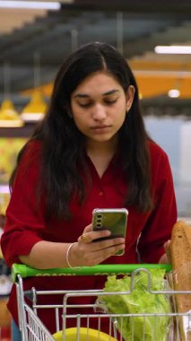 Taze ürünlerle dolu bir alışveriş arabasına yaslanan güzel Hintli kadın, mağazadaki alışveriş listesini kontrol etmek için akıllı telefonunu kullanıyor.