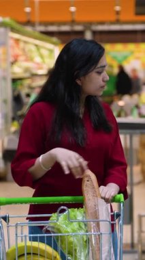 Genç Hintli bir kadın süpermarket koridorunda alışveriş arabasını itiyor. Taze ekmek, marul, muz ve paketlenmiş somon alıyor.