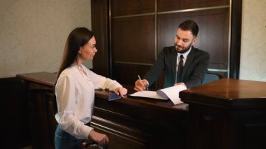 Bir otel resepsiyonunda bavulu olan bir bayan yolcu. Bir erkek resepsiyonist ona kayıt ve evrak işlerinde yardım ediyor.