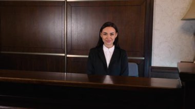 Kendine güvenen ve güzel bir otel yöneticisi klasik bir ahşap resepsiyonda oturuyor. Kolları çaprazlanmış, kameraya gülümsüyor.