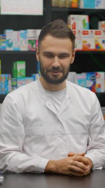 Kameraya gülümseyen sakallı, arkadaş canlısı bir eczacı. Eczanede beyaz önlüklü profesyonel bir sağlık görevlisi.